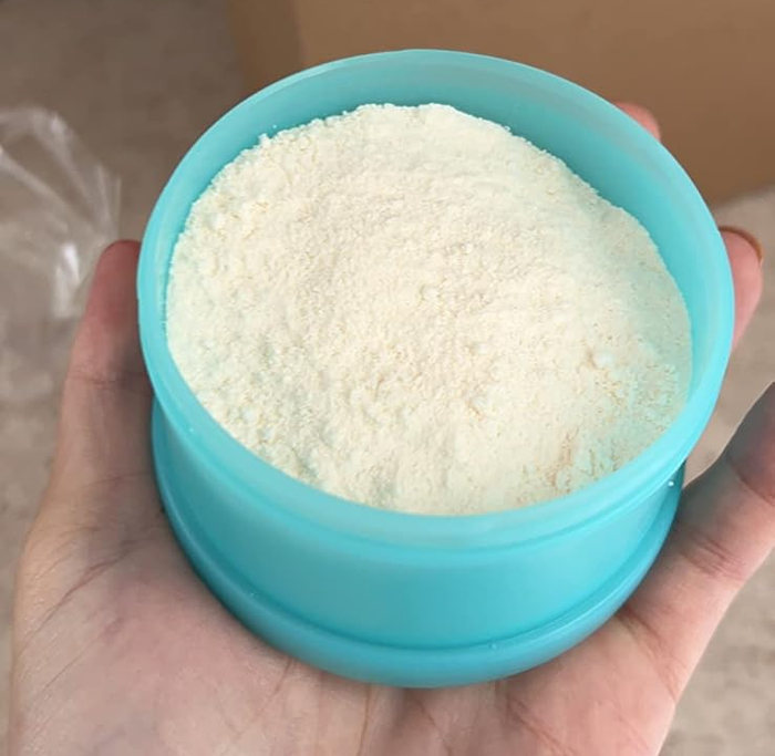 A person holding a blue container filled with a fine white powder, with a partially opened brown box in the background.