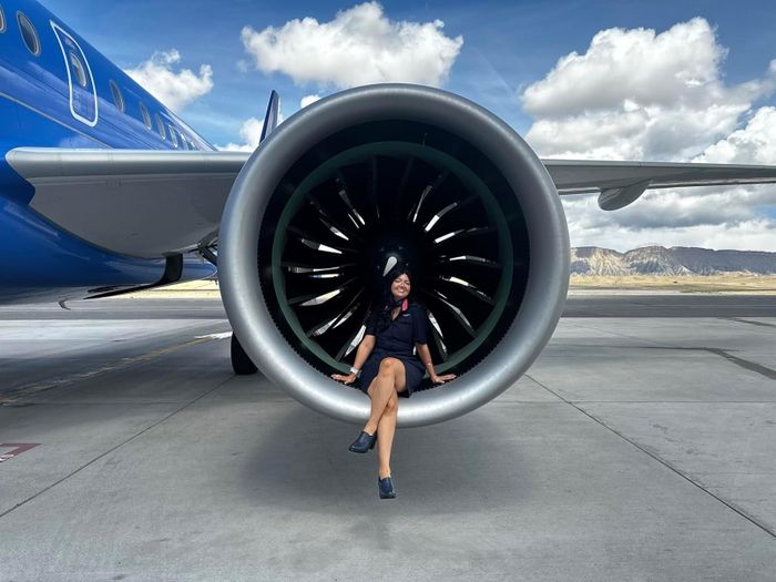 A reviewer, dressed in a flight attendant uniform, poses next to an airplane engine on a runway with a scenic mountain view in the background.