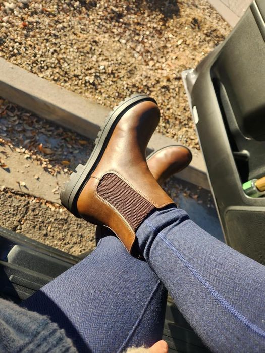 A reviewer wearing brown ankle boots and blue jeans, sitting inside a car with the door open. Leaves and a curb are visible in the background