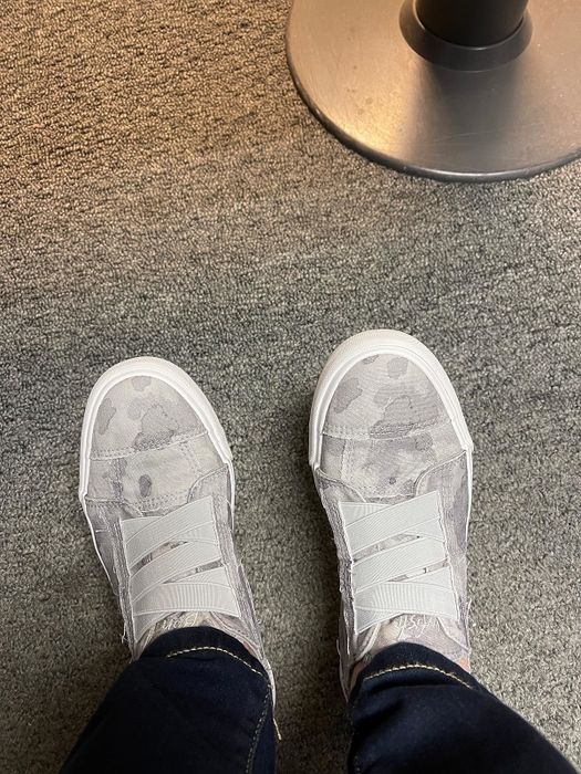 A reviewer shows off camo shoes with criss-cross elastic bands, paired with jeans on a carpeted floor