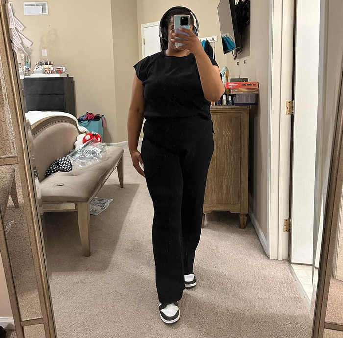 Person snapping a mirror selfie in a black top and pants, paired with sneakers, with a cozy room in the background.
