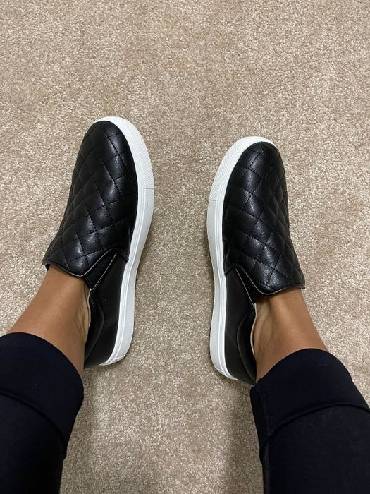 A reviewer casually wearing quilted black slip-on sneakers with white soles, styled with black pants, and relaxing on beige carpet.