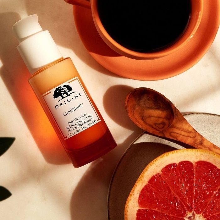A bottle of serum, a sliced orange, a wooden spoon, and a plate with a cup of coffee