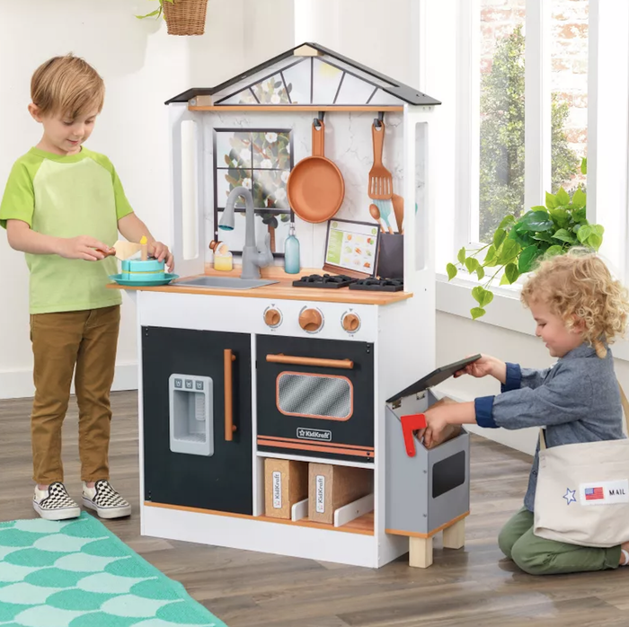 Two kids enjoying their time with a toy kitchen set—one washing dishes at the sink while the other explores the oven.