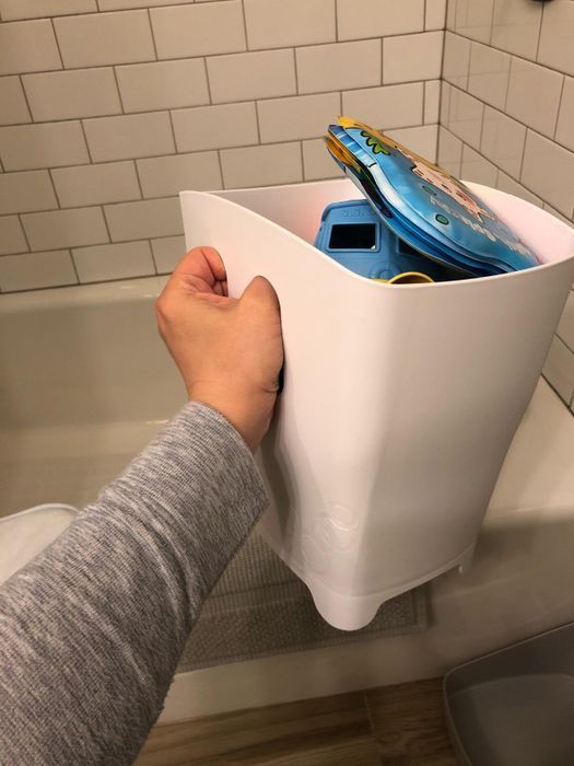 A person holding a white storage container full of bath toys above a bathtub
