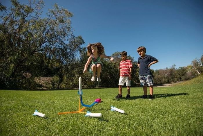 A girl excitedly jumps on a stomp rocket launcher, sending a foam rocket soaring into the sky, while two boys eagerly watch in a grassy field.
