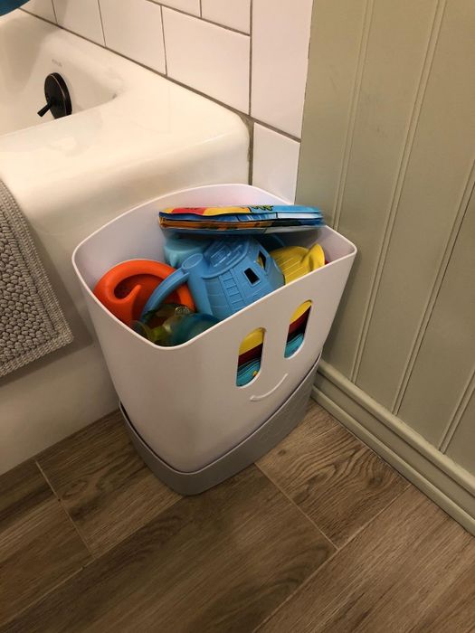 A plastic bin filled with various children's bath toys placed next to a bathtub in a bathroom