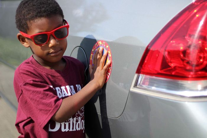 A young child with short hair, wearing red sunglasses and a maroon t-shirt, standing next to a car, with their hand on a decorative magnetic sticker