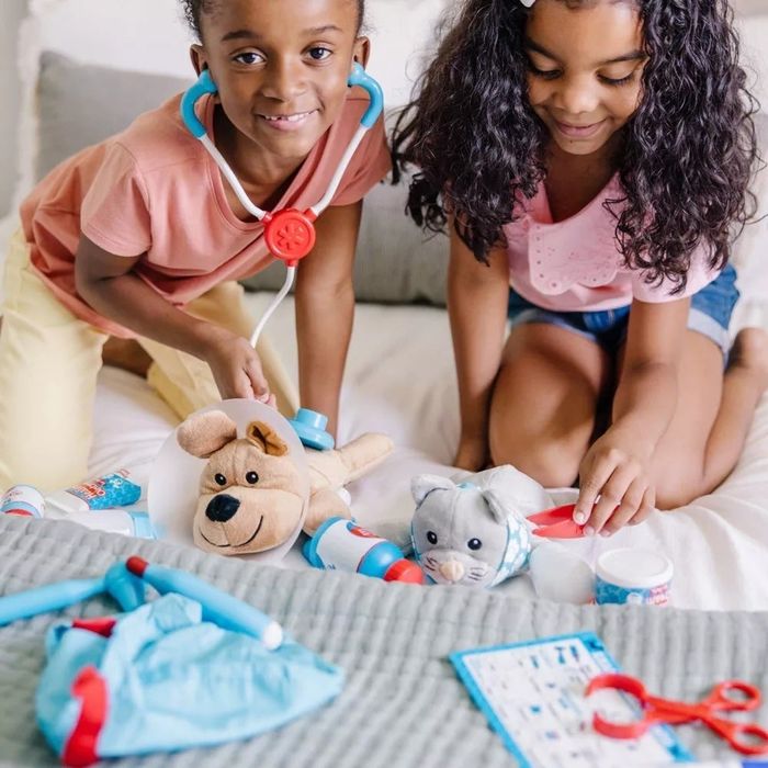 Two young girls happily engage in imaginative play, using a toy doctor kit to treat their stuffed animals on a bed. Their smiles show their excitement as they care for their furry friends.