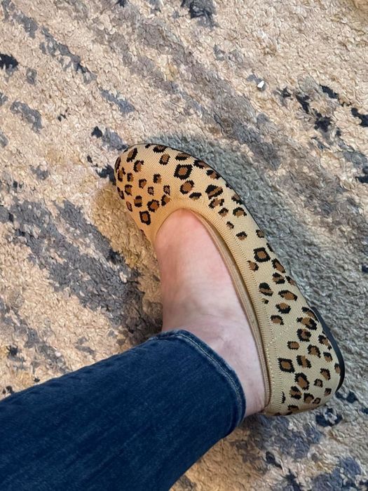 A reviewer’s foot in a leopard print flat shoe, partially visible with denim jeans, resting on a textured rug.
