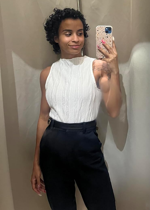 A person trying on a white sleeveless top with black trousers in a fitting room while taking a mirror selfie.