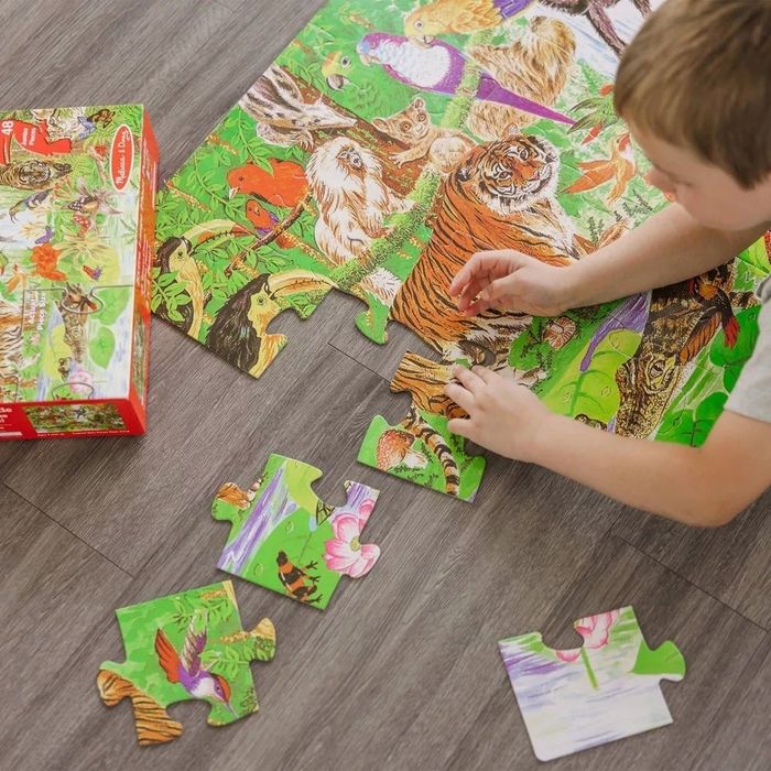 A child putting together a colorful animal-themed puzzle on a wooden floor. Some pieces are still missing, with the puzzle box visible nearby.