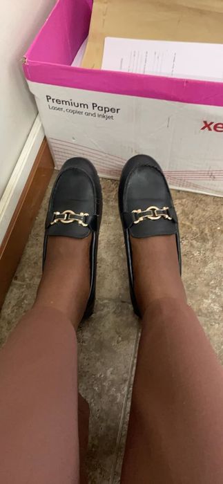 A reviewer sporting black loafers adorned with gold chain details, standing on a tiled floor next to a box labeled 
