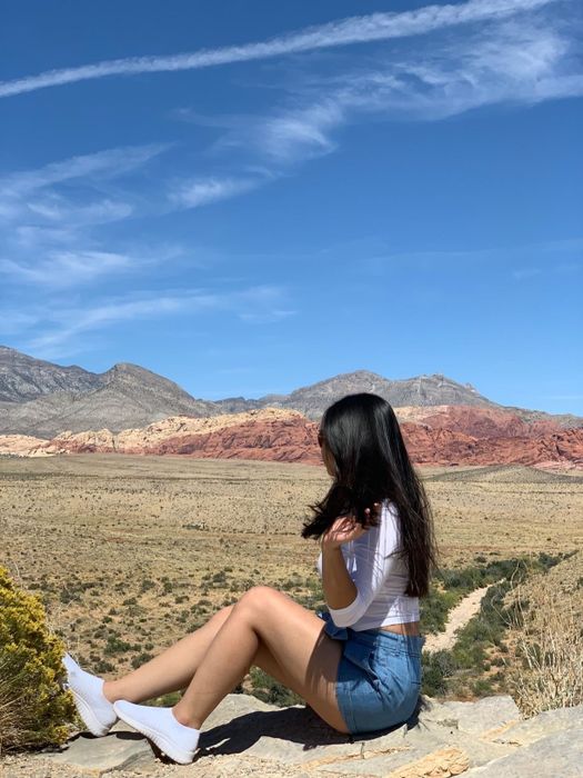 A reviewer dressed in a white top and denim shorts, sitting on a rock and gazing at a desert landscape with mountains in the distance.