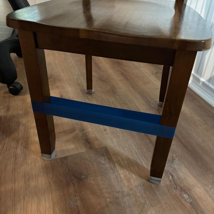 A wooden chair with a blue resistance band wrapped around its legs, placed on a wooden floor in a white-paneled room