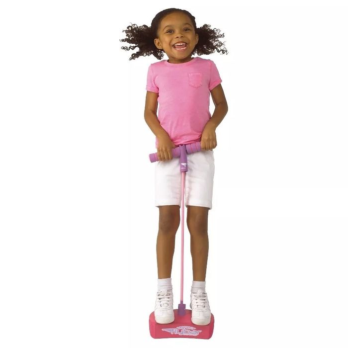 A young girl joyfully jumps on a pink pogo stick, wearing a pink shirt and white shorts, enjoying her time outdoors.