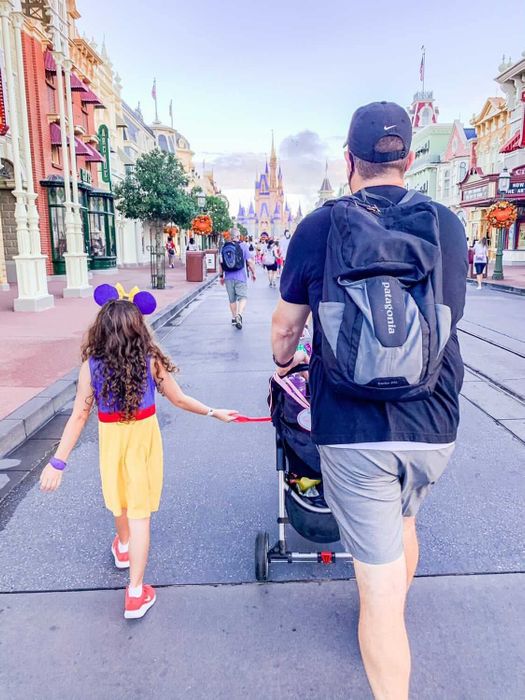 A person in casual clothes pushes a stroller down Main Street, U.S.A., at Disney World, while a young child dressed as Snow White with Mickey ears walks beside the stroller, holding onto a strap attached to it