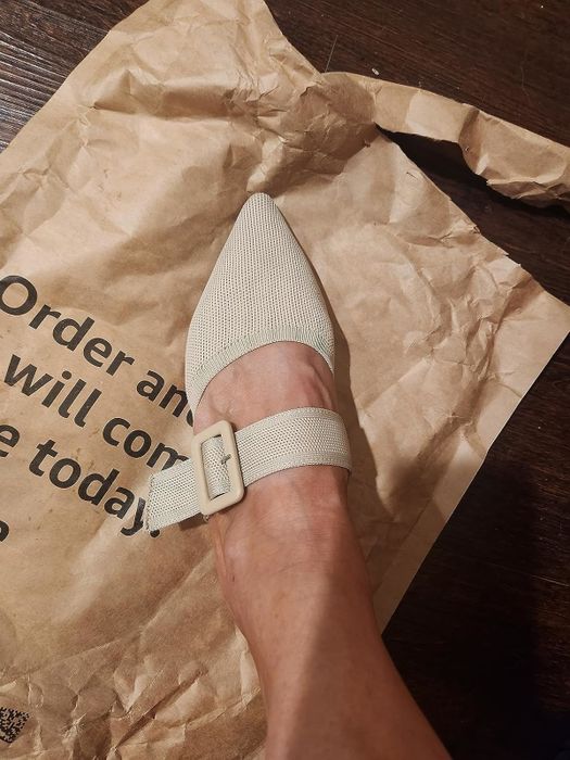 Foot wearing a pointed-toe shoe with a buckle strap resting on a crumpled paper bag, with partial text visible.