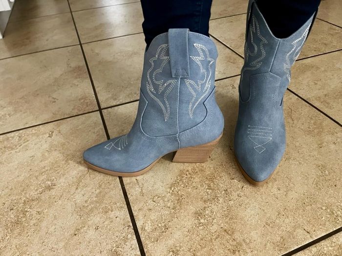 A reviewer showcasing blue cowboy boots with intricate stitching, worn indoors on a tiled floor