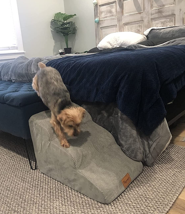 A side-view image from the reviewer of the product placed at the edge of a bed with a dog walking down the stairs on a gray carpet