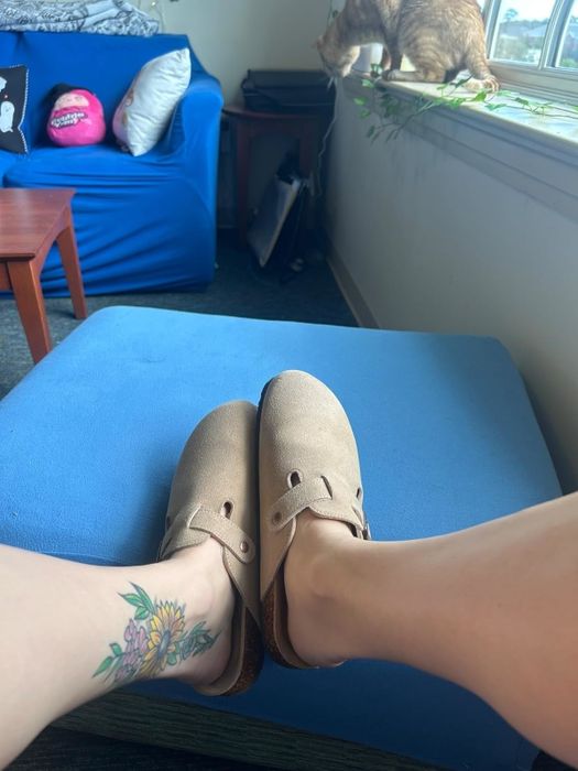 A reviewer with a floral tattoo relaxing in slip-on shoes, legs stretched out on a blue ottoman, with a cat lounging by the window in the background.