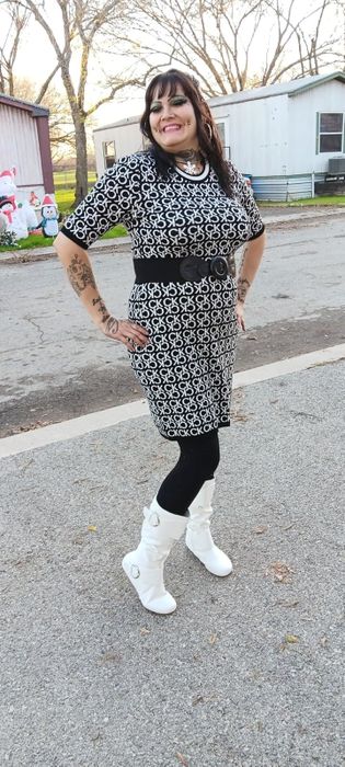 A reviewer wearing a patterned dress and white boots poses outdoors with a smile