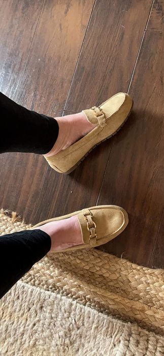 A reviewer in tan suede loafers with a chain accent, standing on a wooden floor