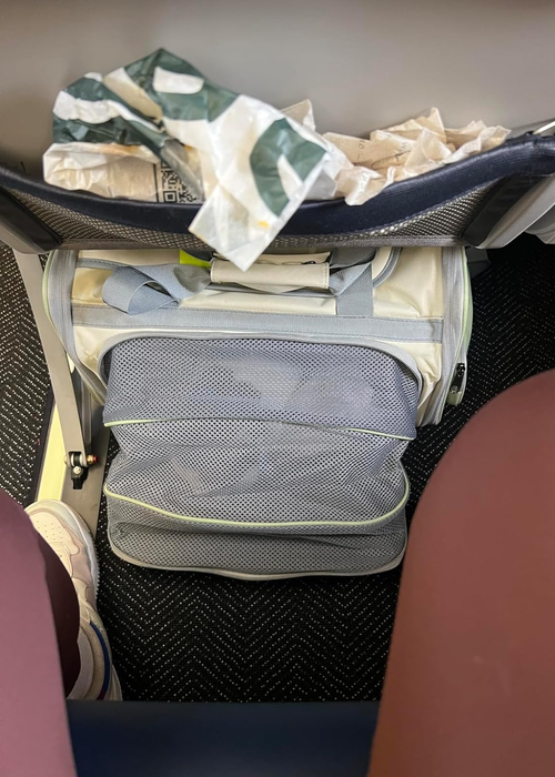 An open suitcase under an airplane seat, filled with clothes and some scattered trash — a perfect example for illustrating packing tips