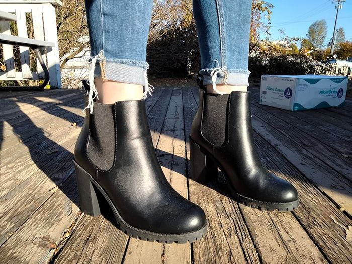 Close-up of a reviewer wearing black leather ankle boots with block heels and rolled-up jeans, standing on a wooden deck