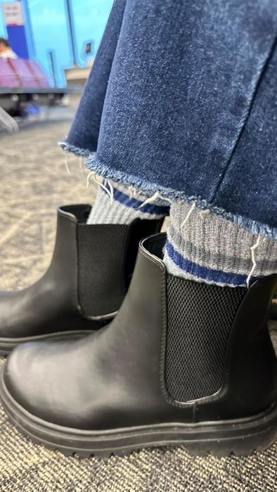 A close-up of a reviewer sporting frayed jeans, gray socks with blue stripes, and black Chelsea boots. A chrome-themed shopping setting is visible in the background
