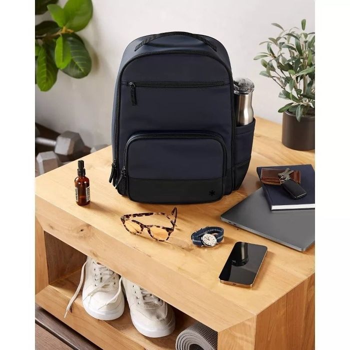 A backpack placed on a wooden desk, surrounded by everyday items like a water bottle, glasses, sneakers, phone, notebook, and a plant, offering a clean, organized look.