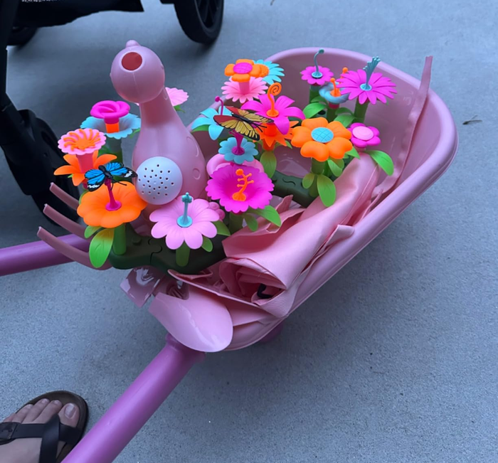 A pink toy wheelbarrow with vibrant play flowers and a watering can resting on a patio