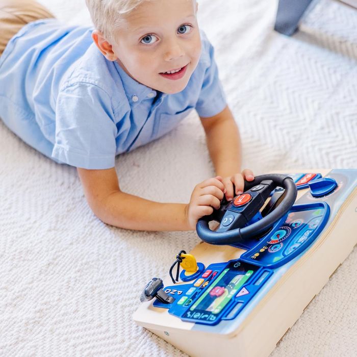 A young child lays on the floor, engaged with a toy car dashboard featuring various buttons and a steering wheel