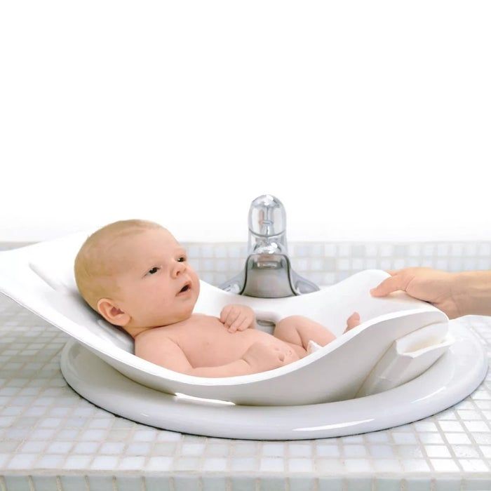 Infant resting in a bath support inside a sink, with an adult's hand gently guiding the side