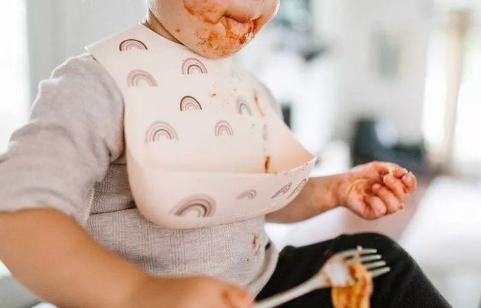 Toddler with food on their face, holding a fork and wearing a rainbow-patterned bib in a high chair