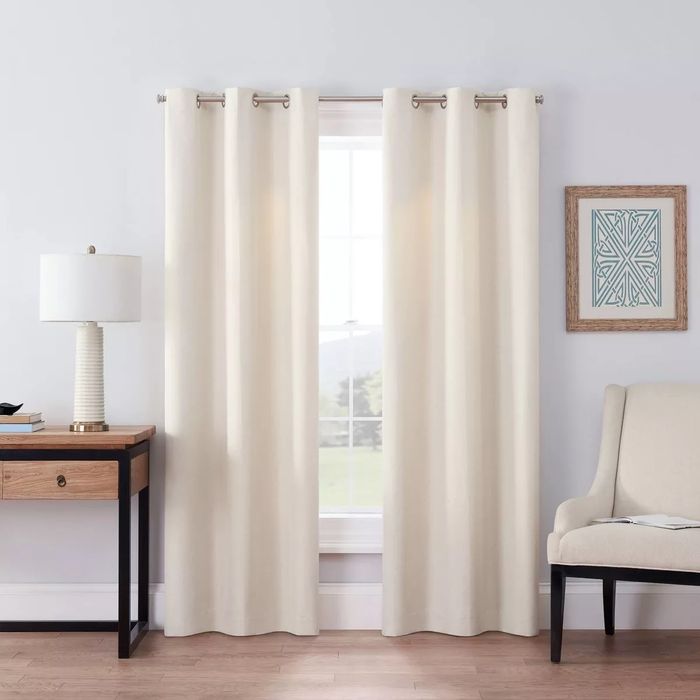 Cream-toned curtains hanging next to a beige armchair and a wooden side table with a lamp.