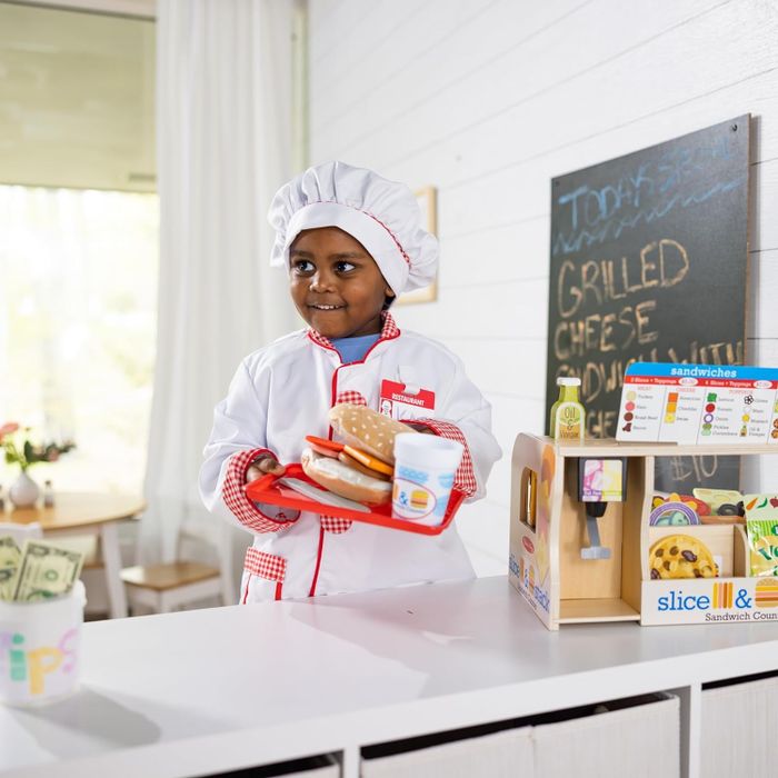 A young child dressed as a chef presents a toy sandwich, smoothie, and a tray at a pretend kitchen station with a toy cash register
