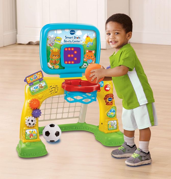 A young child enjoys playing with the VTech Smart Shots Sports Center, holding an orange basketball with a big smile