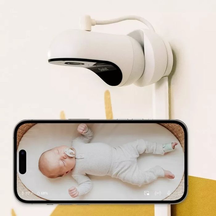 A baby monitor camera positioned above a smartphone displaying a live feed of a peacefully sleeping baby in a onesie.