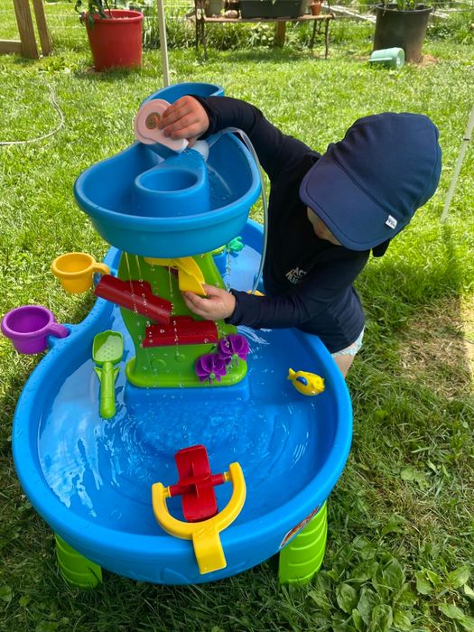 A toddler in a sun hat enjoys playing with a water activity table, complete with various toys and mechanisms like cups, a fishing rod, and a waterwheel