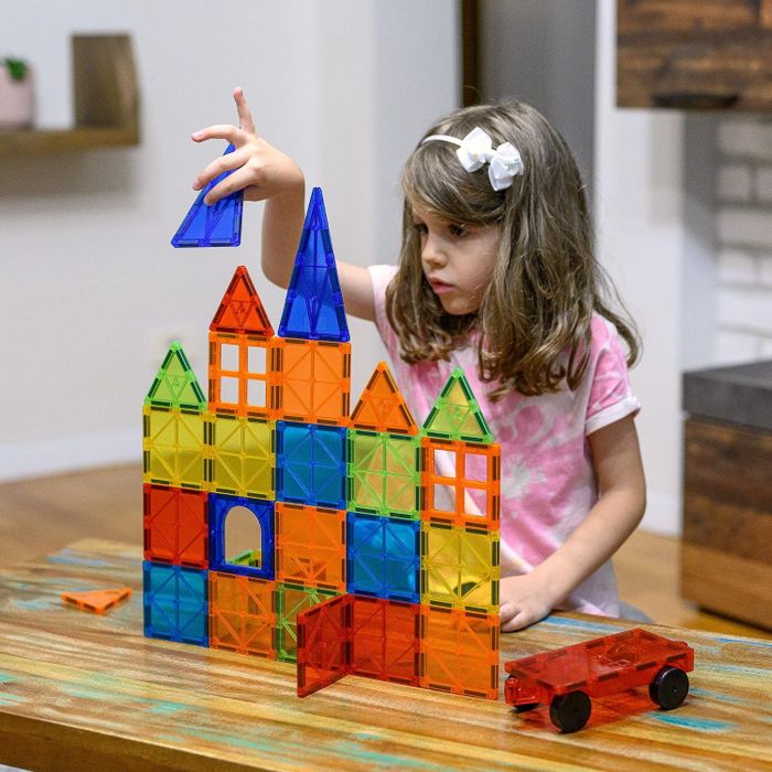 A young child is building a vibrant structure with magnetic tiles, with a toy car resting on a wooden table