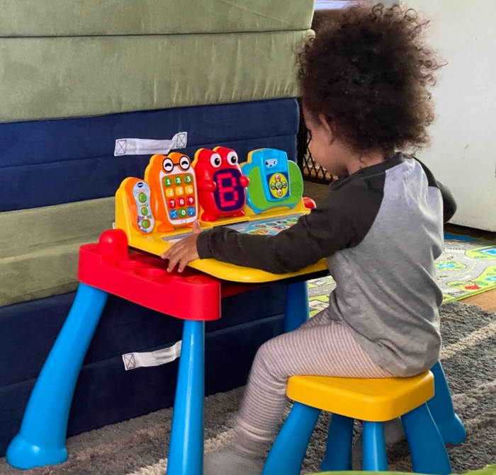 A toddler sits at a vibrant toy desk, interacting with various buttons and educational displays