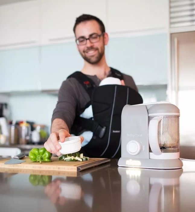 A model cooking in the kitchen with an infant in a carrier, using a countertop baby food processor to prepare homemade meals.