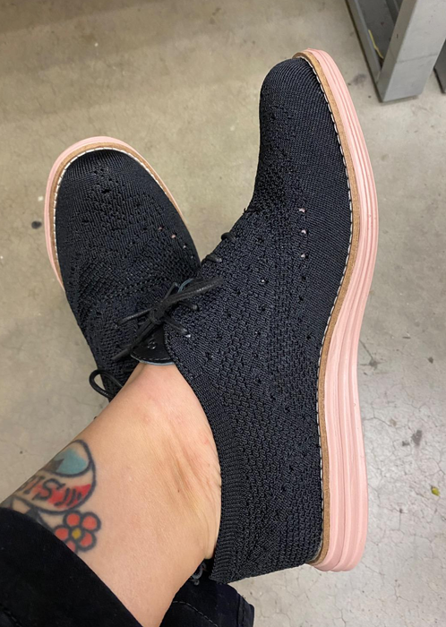 A person's legs featuring a tattoo, wearing black knit-style oxfords with light pink soles