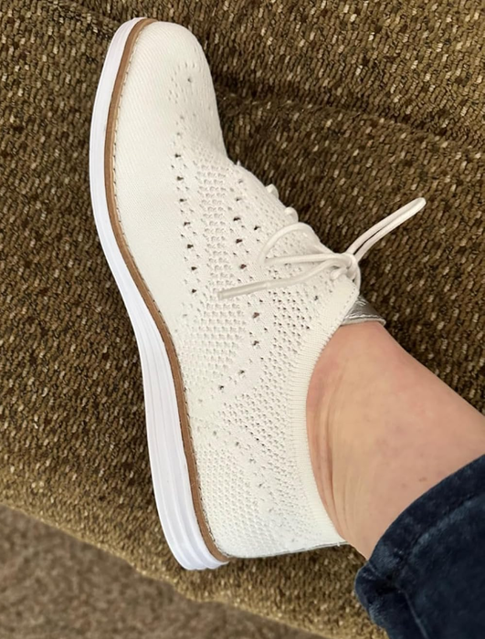 A close-up of someone's foot sporting a white knit lace-up oxford on a textured surface