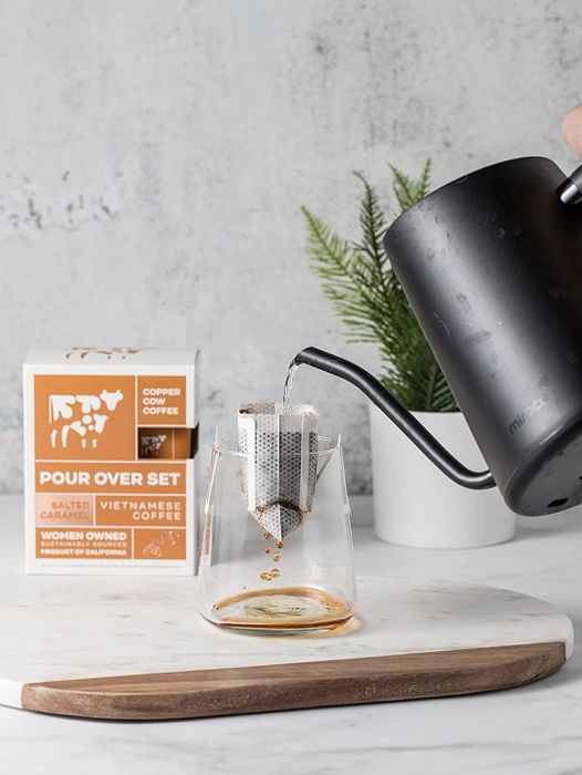 Hand pouring hot water from a kettle over a coffee pod placed on a clear cup, with coffee dripping out from the bottom