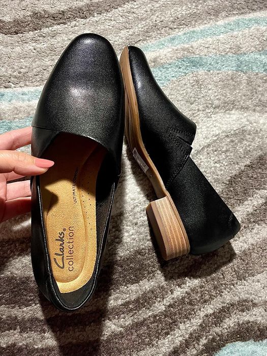 A person holding a pair of black Clarks Collection shoes with a low wooden heel, displayed on a textured surface