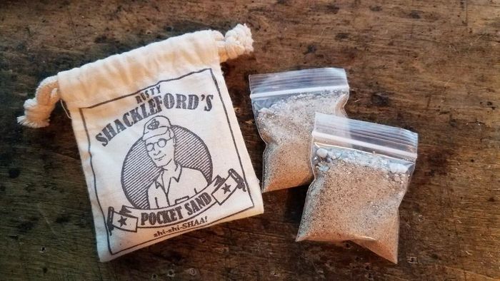 A drawstring bag labeled 'Rusty Shackleford's Pocket Sand,' featuring Dale's face, accompanied by two plastic bags of sand
