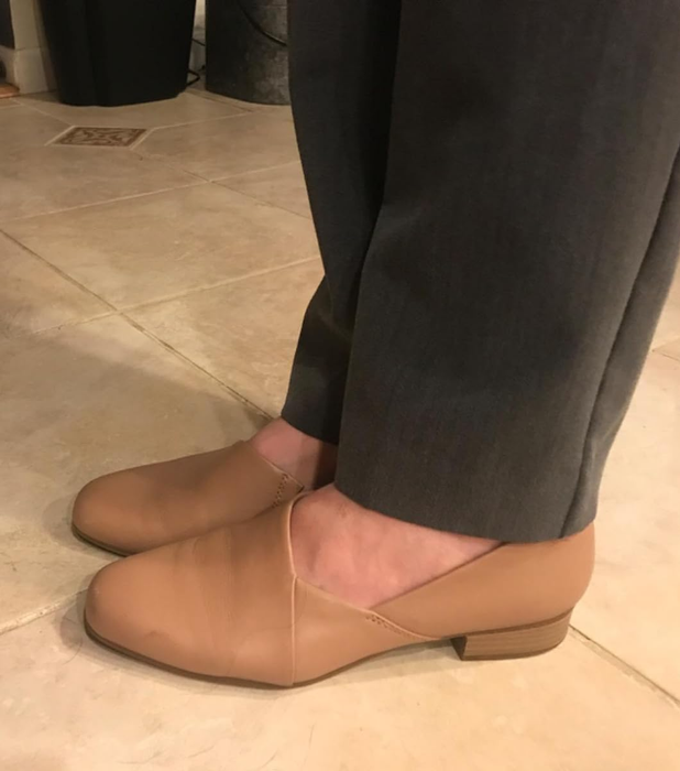 Close-up of someone wearing brown loafer-style shoes with heels and grey trousers, standing on a tiled floor