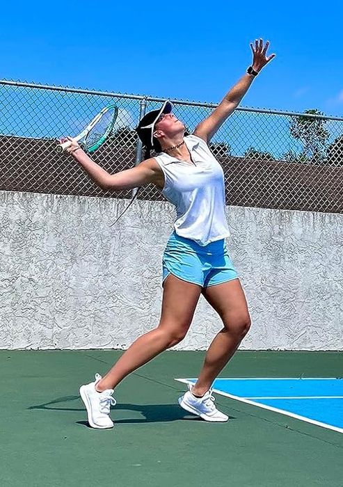 A reviewer playing tennis in a white top, blue shorts, and white sneakers